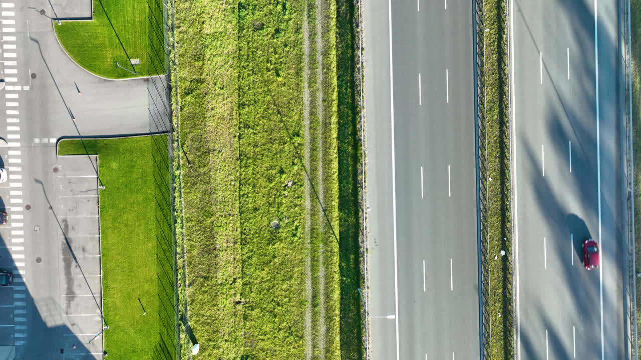 Drone overhead view of parked cars and a green verge aligned beside a highway with a moving vehicle casting a long shadow