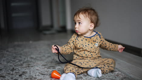 lovely boy sitting on the carpet at home holding a shoe lace. black domestic cat jumping near the kid and stops looking at child.