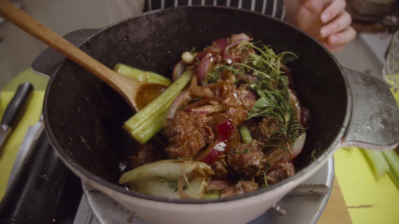 深鍋の中で、牛すね肉、薄切り玉ねぎ、セロリ、スパイスを木べらで調理し、かき混ぜる
