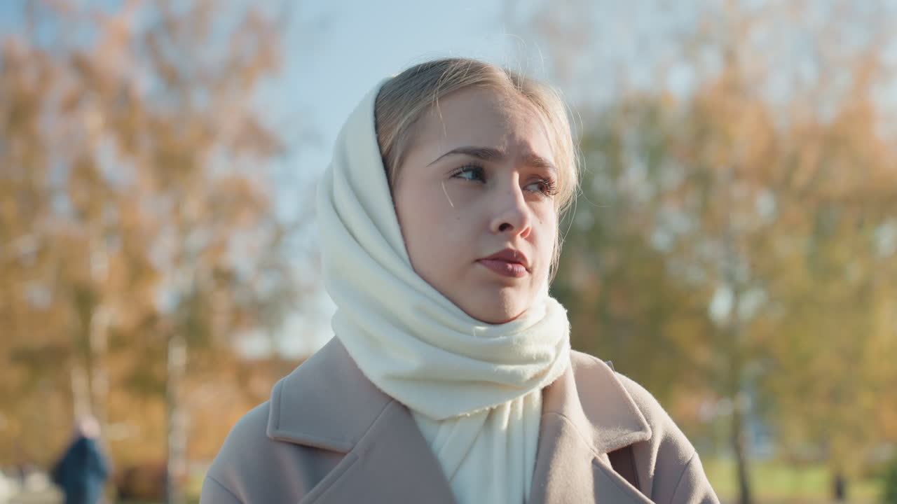 Giovane donna caucasica in sciarpa in una passeggiata contemplativa al parco. Alberi autunnali e luce solare, cappotto beige. Sguardo pensoso verso lo skyline cittadino, atmosfera lenta e cinematografica. Studentessa introspettiva in pausa tra le commissioni.
