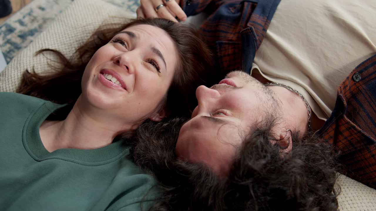 A happy brunette girl in a green jacket lies head to head with a guy with curly hair and talks and laughs while lying on the bed at home