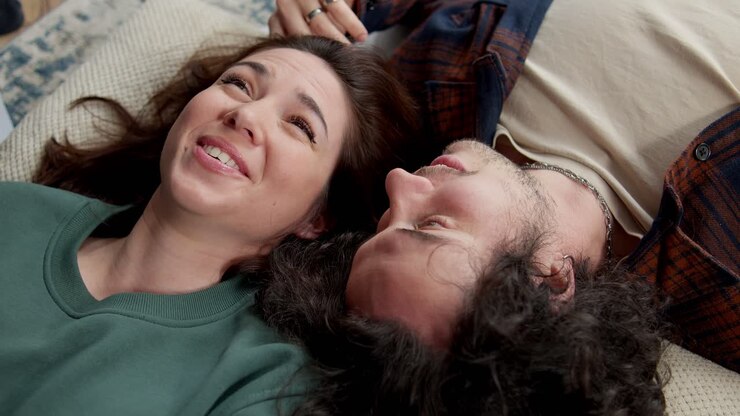 A happy brunette girl in a green jacket lies head to head with a guy with curly hair and talks and laughs while lying on the bed at home