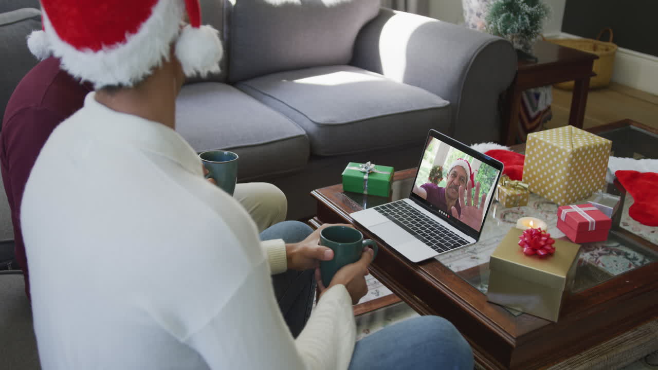 padre y hijo biraciales con sombreros de santa usando una computadora portátil para una videollamada de navidad con un hombre en la pantalla
