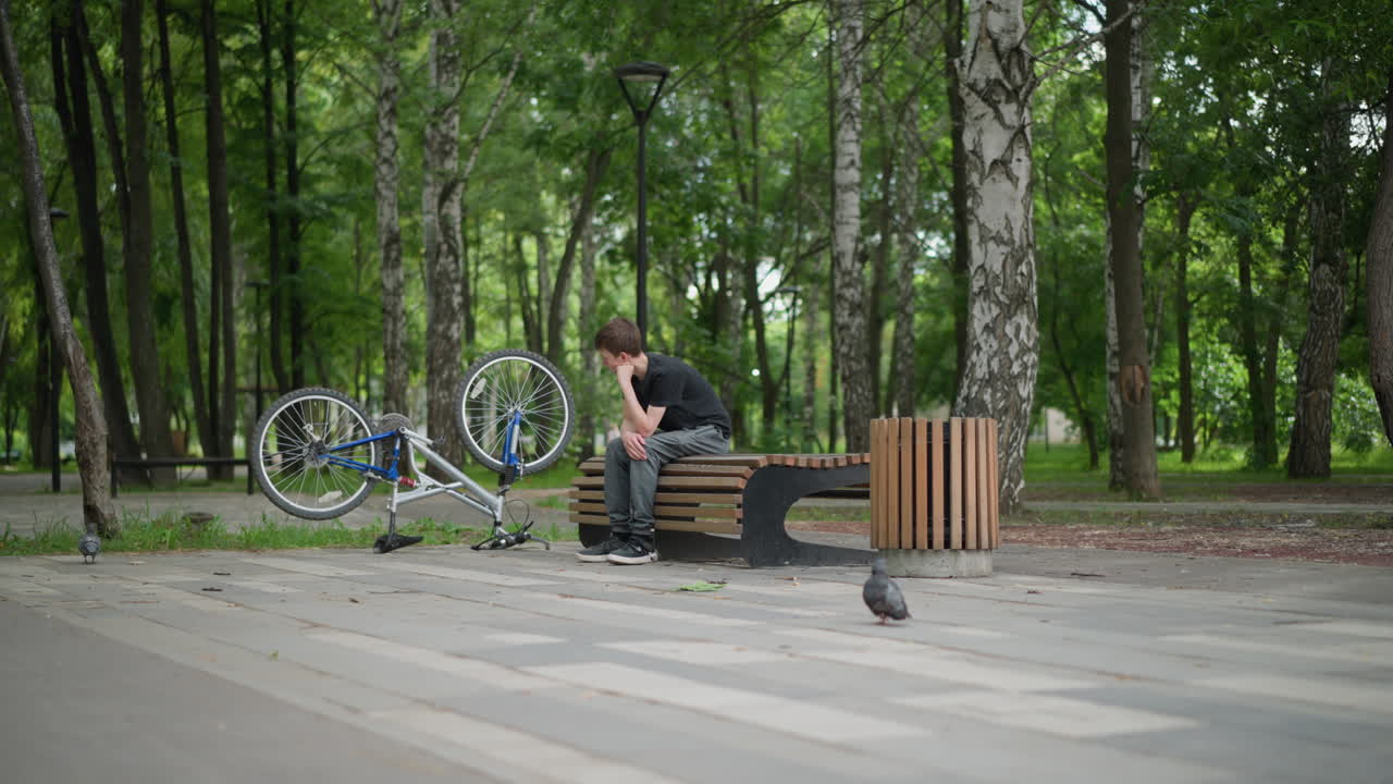 un niño blanco se sienta pensativo en un banco del parque, su mano descansando bajo su barbilla, a su lado hay una bicicleta al revés, en el fondo, con una vista ligeramente borrosa de palomas caminando por el suelo del parque