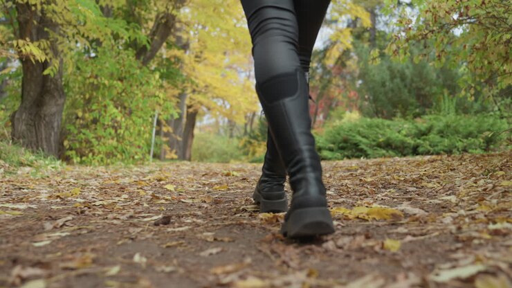 boots walking on fallen leaves along dirt trail, leather leggings and sturdy soles, purposeful energetic stride, close low perspective showing tread imprint and leaf crunch, crisp autumn light
