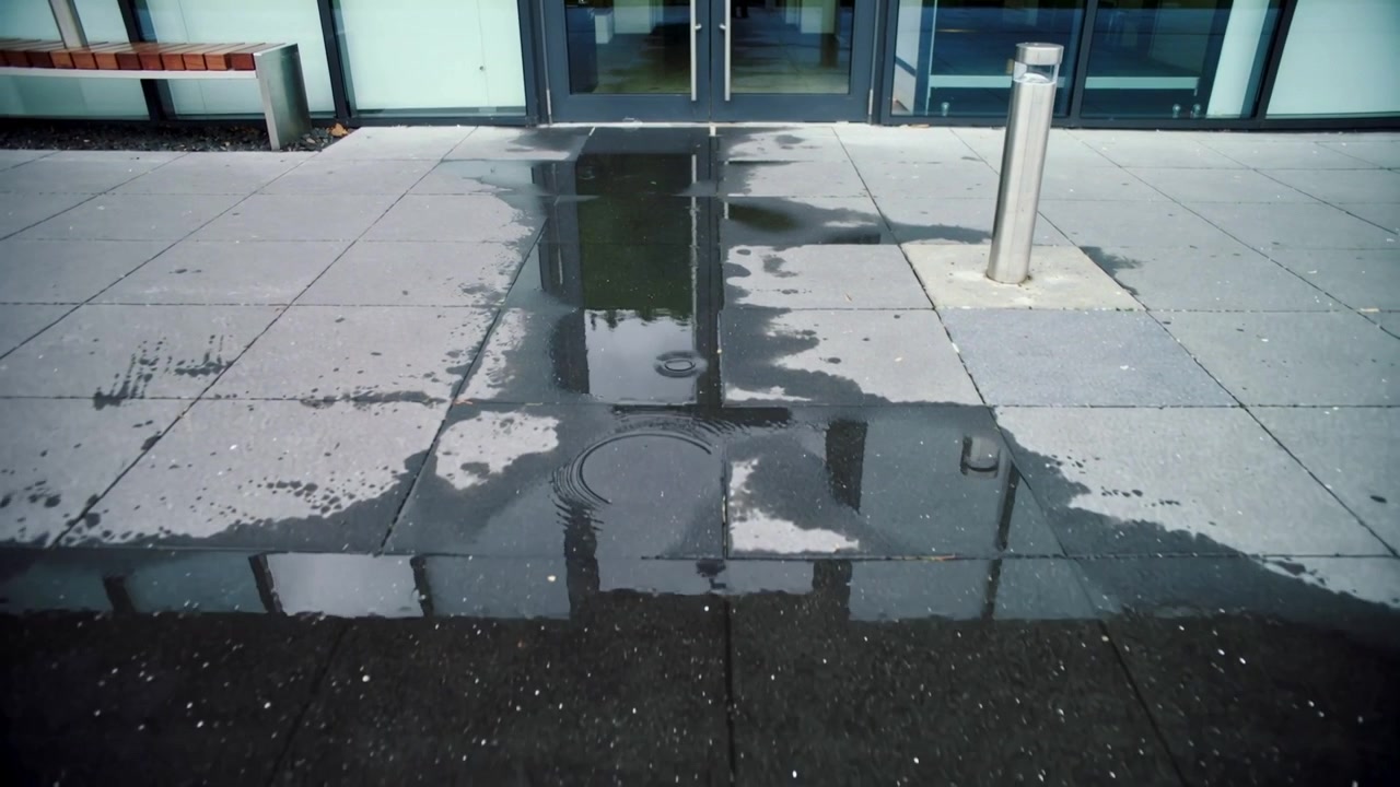 Charco poco profundo con ondas que refleja puertas de cristal en la entrada de un edificio, la lluvia crea anillos concéntricos