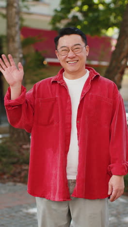 asian man smiling friendly at camera, waving hands hello, hi, greeting or goodbye in city street