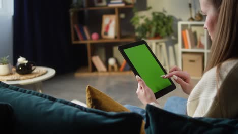 mujer usando una tableta de gadget con pantalla verde. persona femenina irreconocible deslizando la llave de croma, sentada en el sofá en la sala de estar. compras en línea, navegar por internet. tiempo libre en casa, ocio