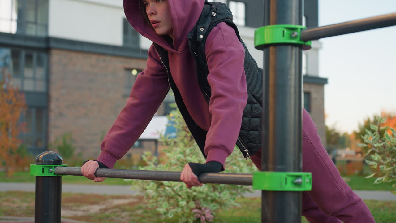 Active woman exercising on outdoor fitness equipment, performing press-ups on horizontal rod in urban park, building strength and endurance. Focused on maintaining proper form during workout