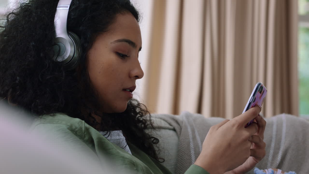 happy teenage girl listening to music wearing headphones relaxing on sofa at home texting with smartphone enjoying sharing lifestyle on social media