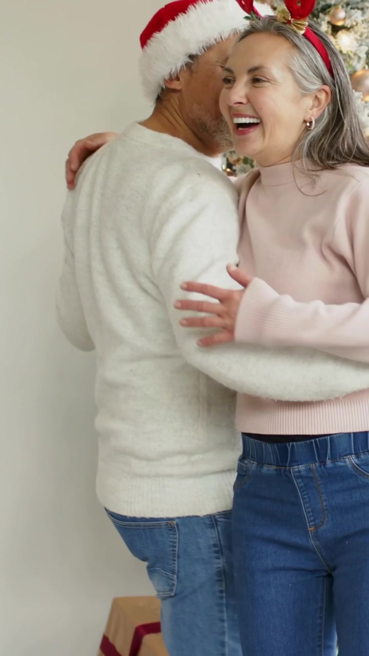 Video verticale: Uomo che cammina con un cappello da Babbo Natale verso una donna con corna che guida persone che ballano con maglioni e jeans vicino all'albero.