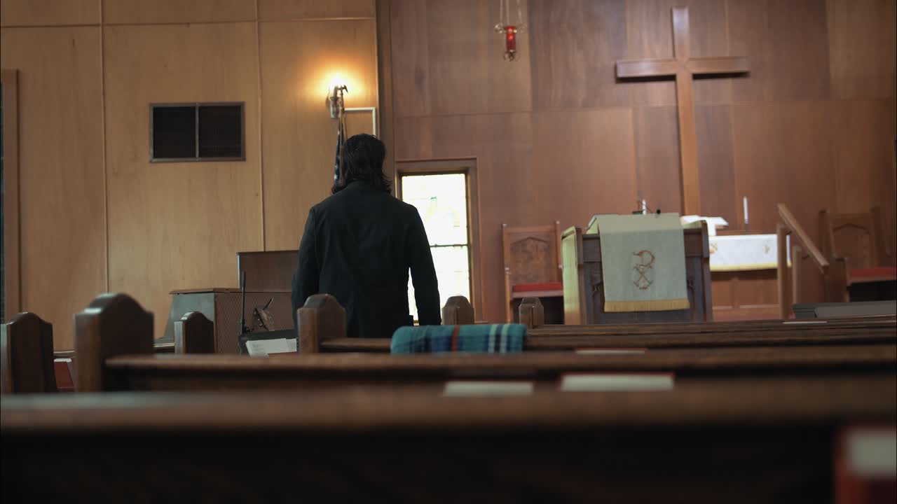 검은색 의상을 입은 기독교 목사가 교회에서 고 영화 속도 느린 동작으로 벤치에 앉아 있습니다.