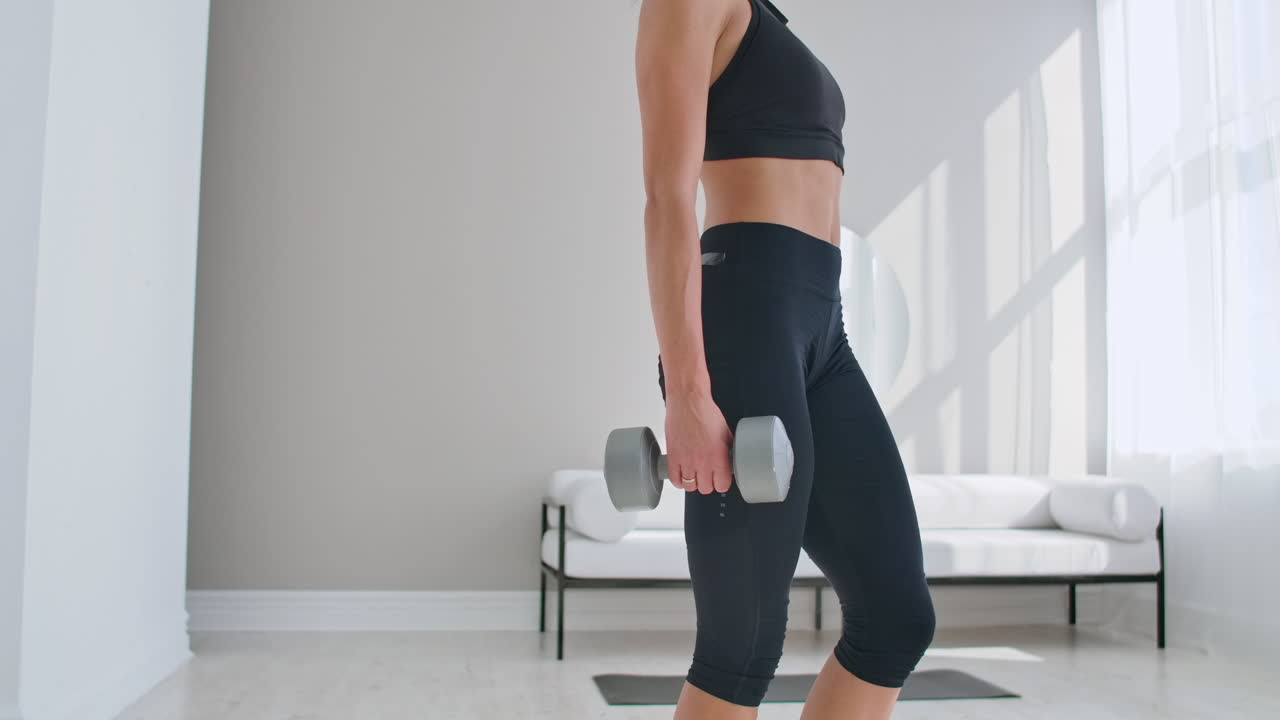 은 날에 집에 있는 여자가 거실에서 손에 덤벨을 들고 뒤로 니다. 건강한 생활 방식 매일 집에서 운동하십시오.