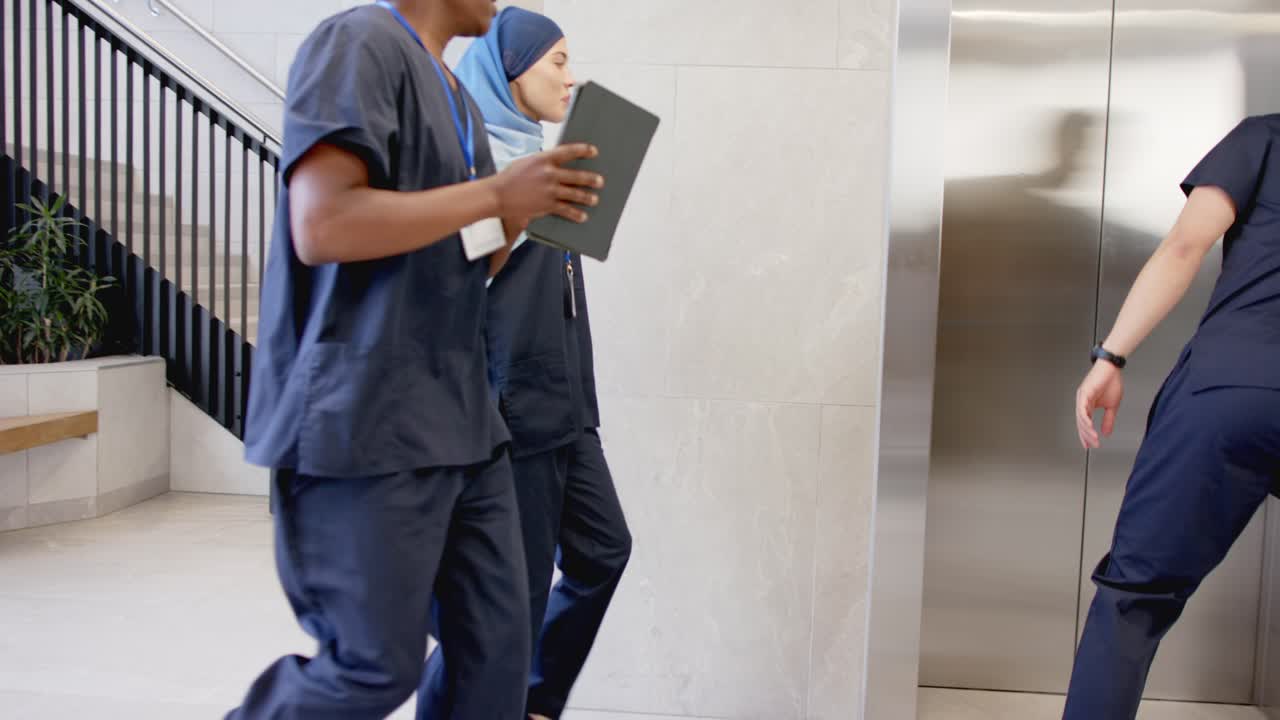 Equipe diversa de scrubs no saguão após o líder asiático apontar para o elevador, pegando um arquivo no tablet para as rondas.
