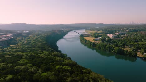 Aerial-tilt-up-over-Lake-Austin-greenbelt-to-reveal-Pennybacker-360-bridge-during-hazy-summer-sunrise-in-Austin,-Texas