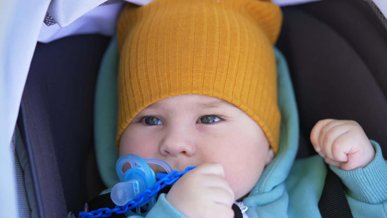 Grey-eyed toddler wearing yellow cap blinking eyes slowly. Lovely boy holding pacifier at his mouth lying in stroller.