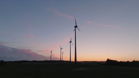 Aerial-establishing-view-wind-turbines-generating-renewable-energy-in-a-wind-farm,-evening-after-the-sunset-golden-hour,-countryside-landscape,-high-contrast-silhouettes,-drone-shot-moving-forward