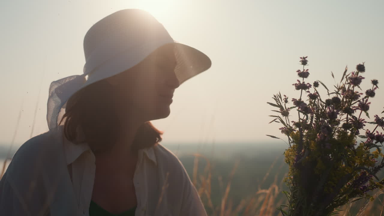 close up of woman in white sunhat gently scenting wildflower bouquet under golden sunlight glow, eyes closed in peace, serene moment in tall grass meadow