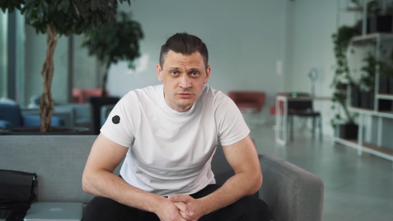 portrait of a young man in a white T-shirt against the backdrop of a business center, sitting on a sofa, adjusting, looking into the camera, and gesturing