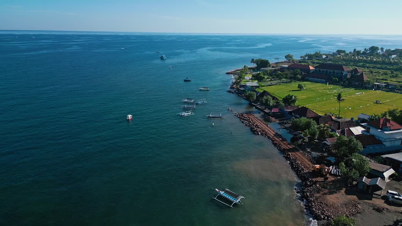 Rekaman udara drone dari garis pantai Bali Utara yang menampilkan ombak lembut, perahu nelayan tradisional, rumah-rumah desa, dan pepohonan tropis yang rimbun di bawah terangnya siang hari di area wisata tepi pantai yang populer
