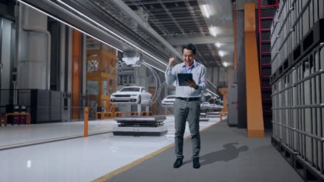cuerpo lleno de un trabajador profesional masculino asiático de pie con su tableta en la fábrica de automóviles ev, instalación industrial de alta tecnología, levanta el puño con el objetivo de gritar después de escribir en la tableta