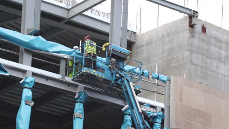 Close-up-of-construction-worker-wearing-protective-clothing-and-safety-harness,-standing-in-the-platform-of-an-elevated-boom-lift,-wrapping-steel-beam-in-a-blue-protective-membrane