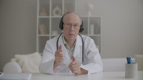 Un-Anciano-Médico-Vestido-Con-Bata-Blanca,-Gafas-Y-Auriculares-Se-Comunica-En-Línea-Con-Un-Retrato-Medio-Frontal