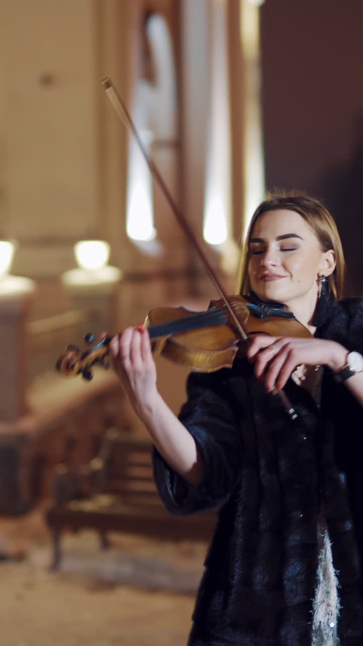 A beautiful girl is playing the violin in the centre of the city in the winter near the road on amid passing public transport. Blurred background. Vertical video