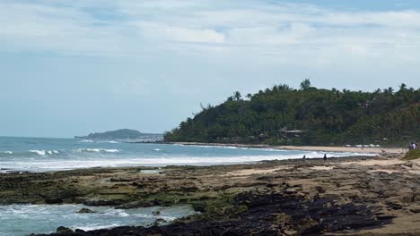 Wide-4k-shot-of-waves-crashing-into-rocks-on-the-beautiful-tropical-Tibau-do-Sul-beach-in-Rio-Grande-do-Norte,-Brazil-covered-in-golden-sand,-palm-trees,-and-cliffs-of-exotic-plants-a-summer-day