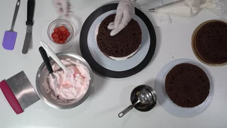 baker placing a new layer of a chocolate cake on top of an already filled layer and proceeding to use a pastry bag to decorate with cream.