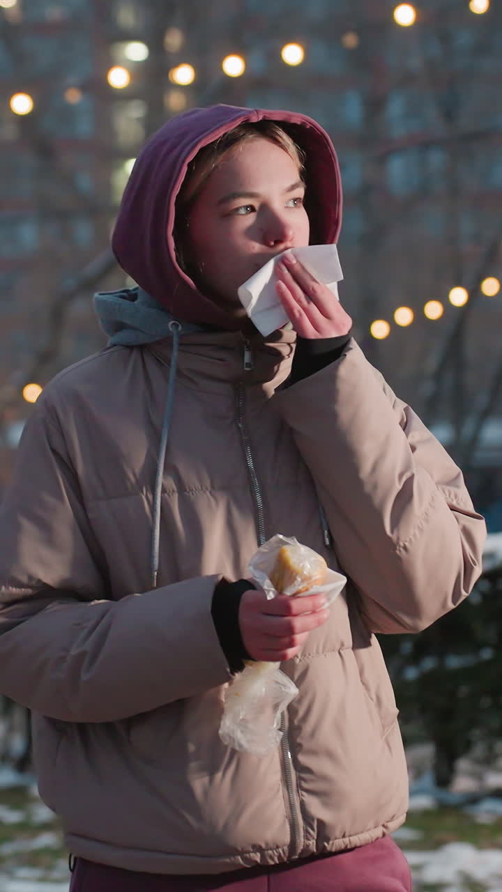 Una donna che tiene uno spuntino in mano e usa un tovagliolo per pulirsi la bocca in un ambiente esterno invernale, con luci bokeh sullo sfondo, mentre si gode il cibo con il freddo in un parco innevato con persone che camminano.