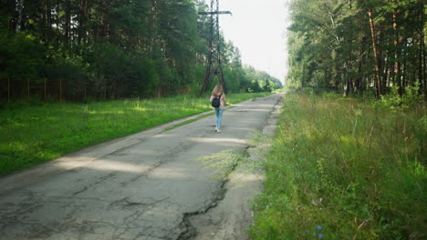 rear view of lady walking alone beside cracked tarred road bordered by lush green forest and tall electric power poles on sunny day, evoking calm, distance, and thoughtful movement in nature