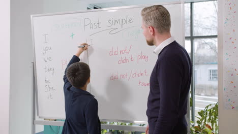 male primary student writing english pronouns on whiteboard while teacher looking at him