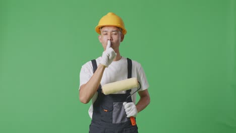 asian man painter wearing safety helmet looking at camera and making shh gesture while standing in the green screen background studio