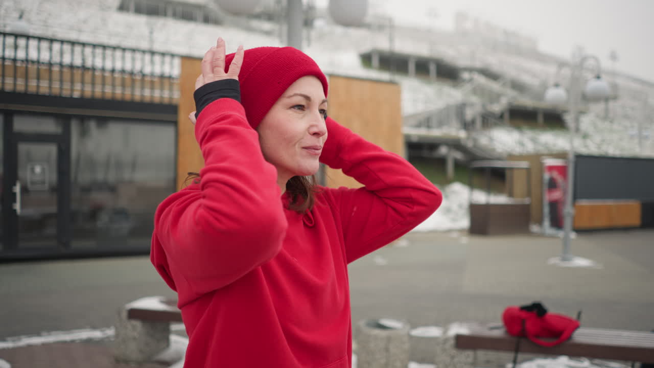vista lateral de un freelance ajustando su gorra roja al aire libre con una cálida sonrisa durante el invierno, el fondo presenta paisajes urbanos cubiertos de nieve, estructuras de madera y bancos modernos