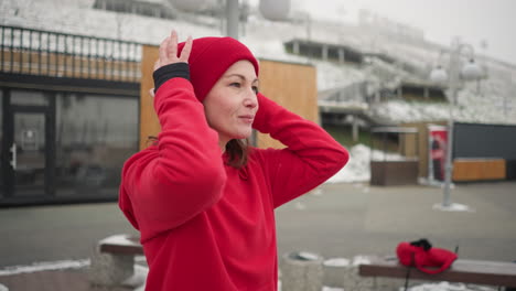 side view of freelancer adjusting red beanie outdoors with a warm smile during winter, background features snow-covered urban landscape, wooden structures, and modern benches