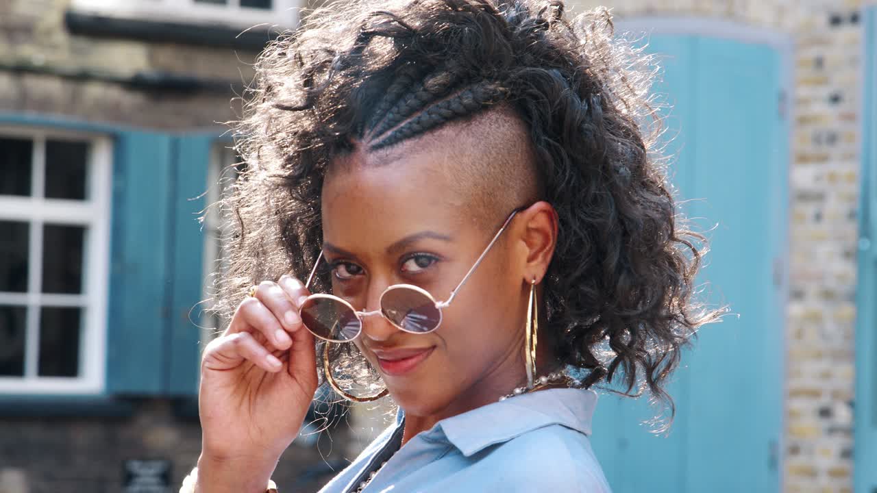 Trendy young black woman in blue dress and sunglasses turns to look to camera, close up, back view