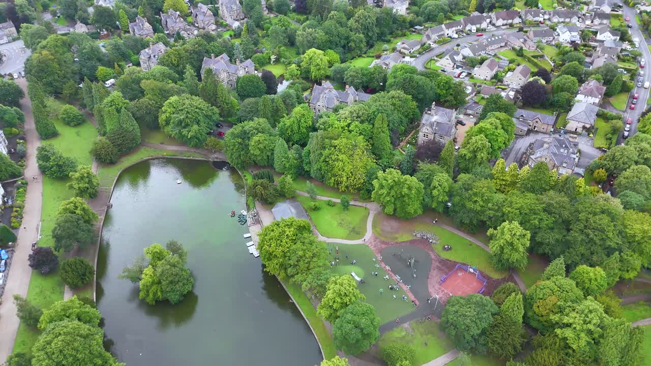Aerial footage glides over a lush green park with a pond, residential buildings, and tree-lined paths under soft natural daylight, creating a tranquil atmosphere