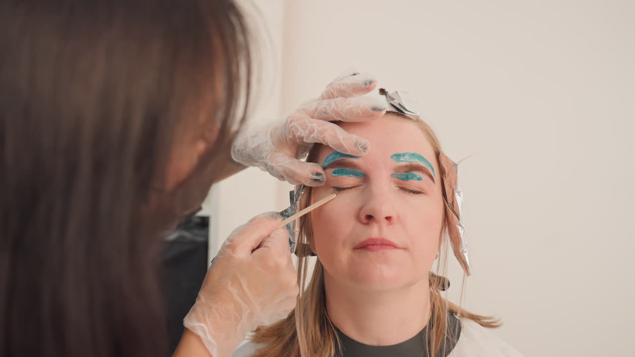caucasian woman receiving eyebrow tinting treatment. esthetician applies blue green pigment with precise brushwork, gloved hands, foilwrapped hair sections, bright minimalist studio, clinical