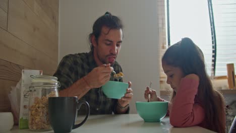 Brunette-man-in-a-green-checkered-shirt,-the-single-father-has-breakfast-with-his-little-daughter,-a-brunette-girl-in-a-pink-dress-at-the-table-in-the-morning-in-a-modern-apartment