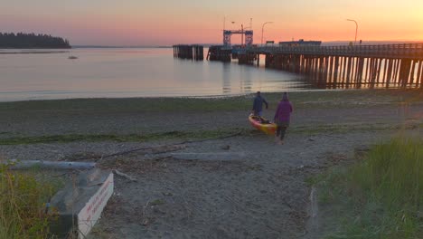 Pareja-De-Hombres-Y-Mujeres-Caminan-Llevando-Kayak-De-Mar-A-La-Bahía-Del-Océano-Al-Amanecer-Cerca-De-Seattle,-Washington