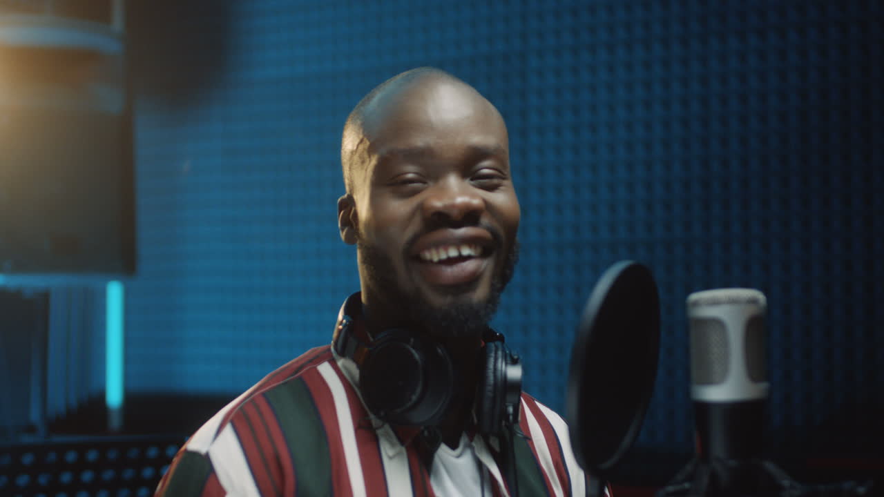 primer plano del joven cantante audaz tomando su sombrero en el estudio de sonido en el micrófono y sonriendo alegremente a la cámara