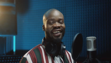 Close-Up-Of-The-Young-Bold-Man-Singer-Taking-On-His-Hat-In-The-Sound-Studio-At-The-Microphone-And-Smiling-To-The-Camera-Cheerfully