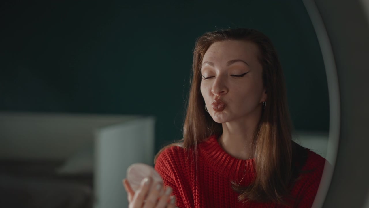 Young woman at vanity applies lipstick before mirror, checks makeup result in compact reflection, then smiles with satisfied expression in soft indoor light during personal beauty routine