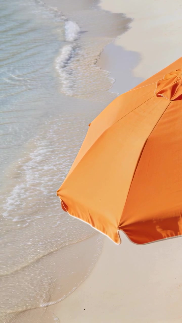 Video verticale: Onde in arrivo che portano schiuma su una spiaggia chiara fino a un ombrellone arancione, con spazio per il testo