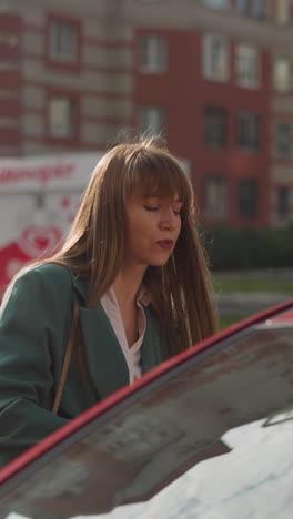 young female driver with long hair gets into red automobile. tired businesswoman rushes home after busy day at work against blurry city buildings