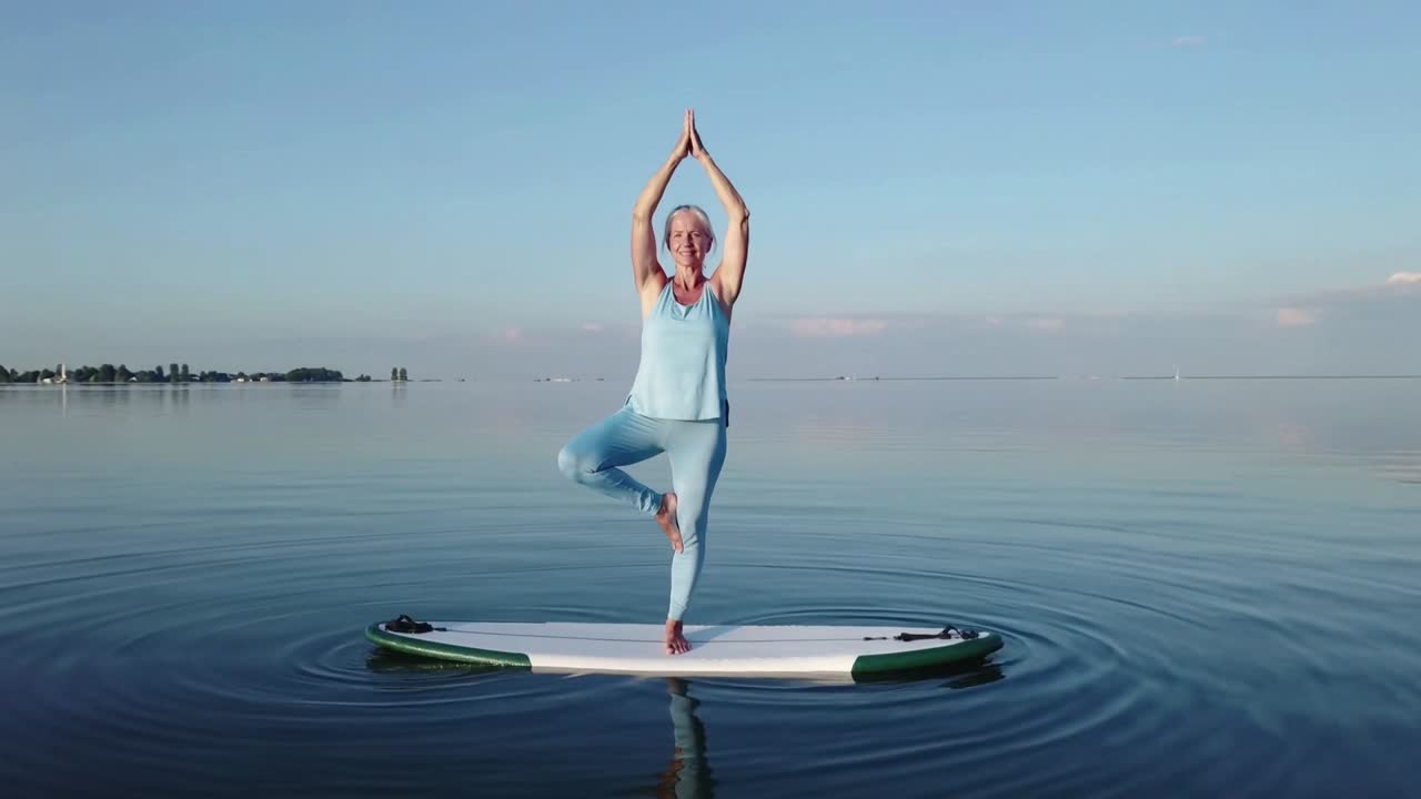 Donna anziana in equilibrio, che mantiene la posizione dell'albero su un paddleboard al lago, indossando abiti da yoga azzurro chiaro.