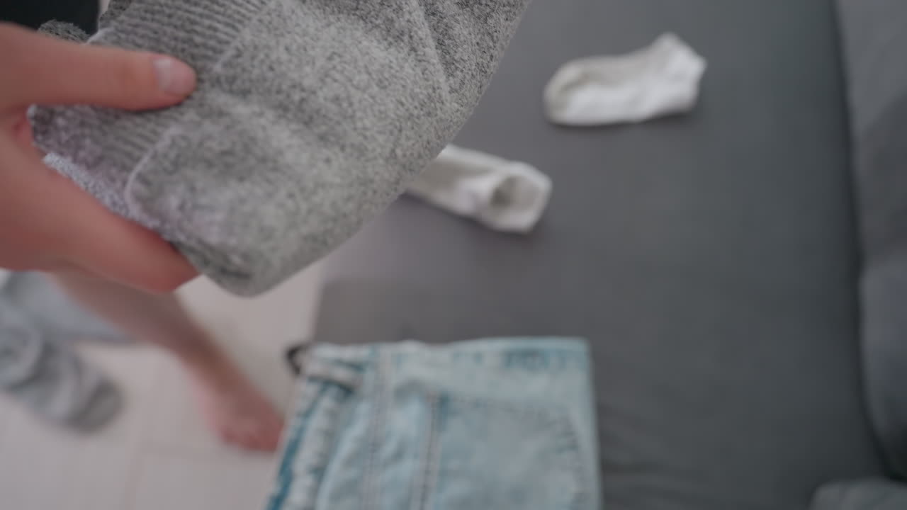 Close up of lady folding gray sweater then dropping it on folded jeans placed on couch showing detail of domestic routine household clothing care lifestyle organization in tidy home environment