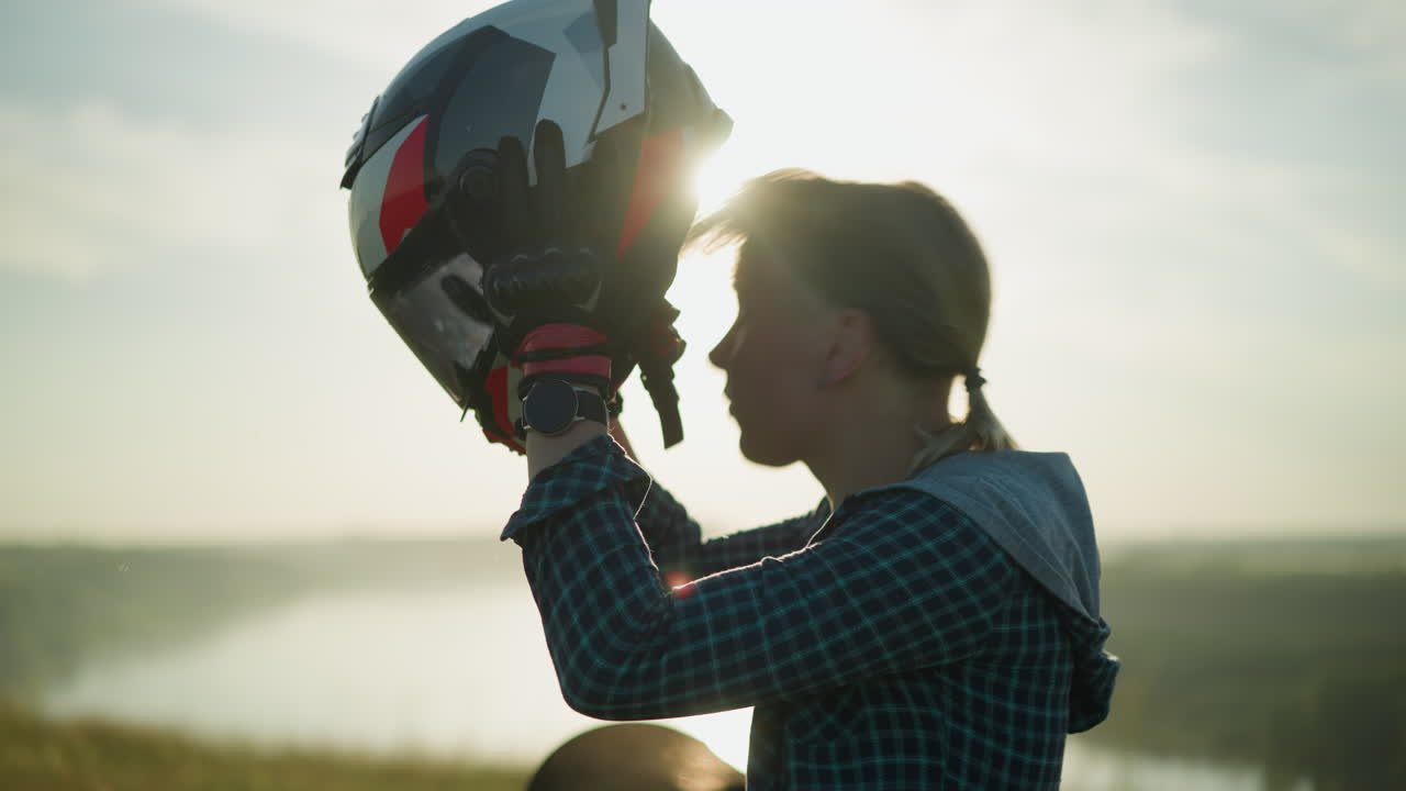 una mujer joven con un casco es capturada en el momento en que se quita el casco, sacudiendo suavemente la cabeza, con la luz del sol iluminándola en el fondo
