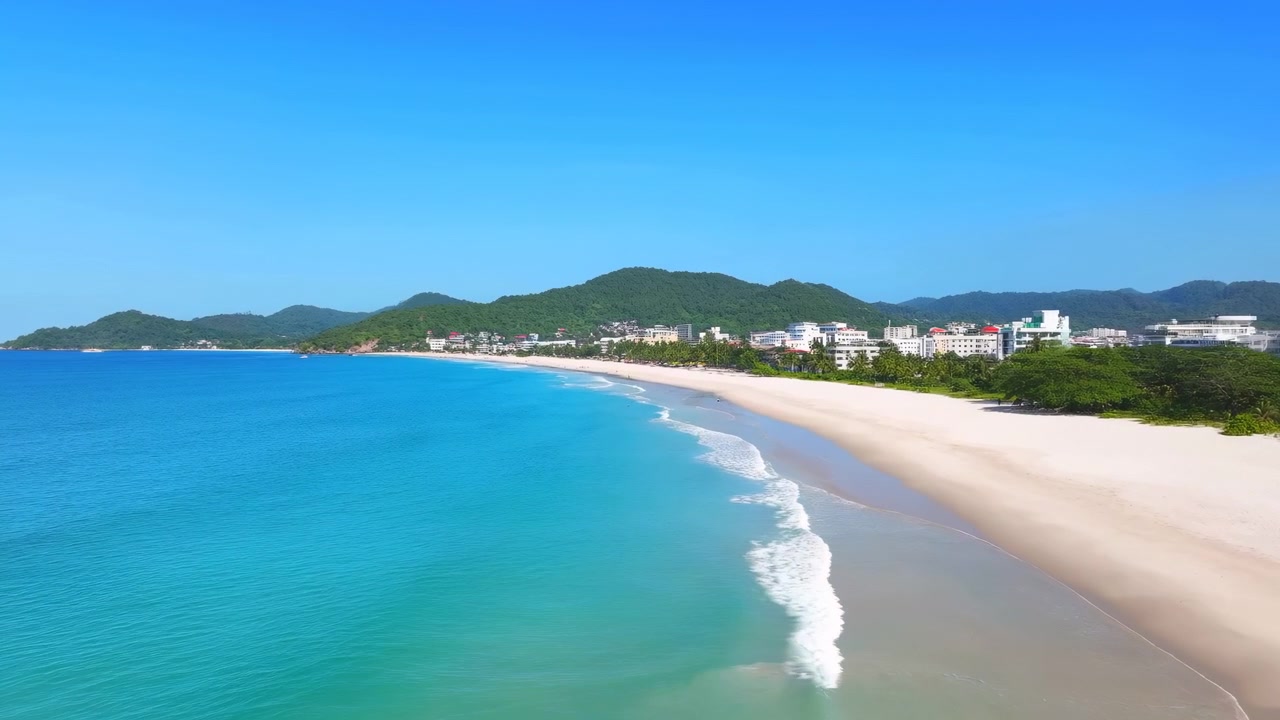 Vista aérea amplia de una costa tropical con agua turquesa, olas suaves, una larga playa de arena blanca, colinas verdes y edificios bajos a lo largo de la costa bajo un cielo azul claro.